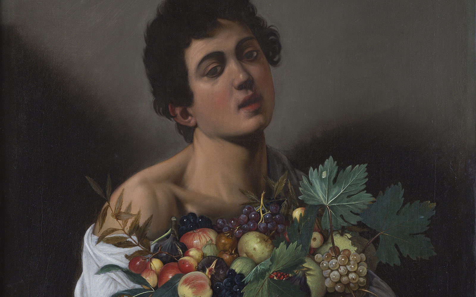 Portrait of a young male with dark hair holding a basket containing grapes, apples, and leaves.