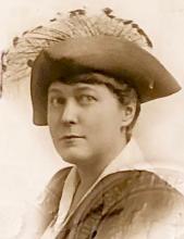 A sepia tinted portrait of a woman looking directly at the camera wearing a tricorn hat with feathers on the top. Her hair appears short in the picture and is a dark color. She wears a white blouse with a dark jacket over it. She is only visible from the shoulders up.