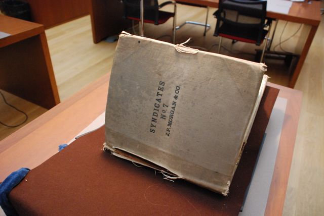A large closed volume, focused on the cover of the book at an angle on a book stand covered in brown felt. The cover of the book is an aged discolored off white with rips on the binding, spine and edge of the book. There are three lines of black text on the front of the book. The text reads Syndicates, No. 7, J.P. Morgan & Co.