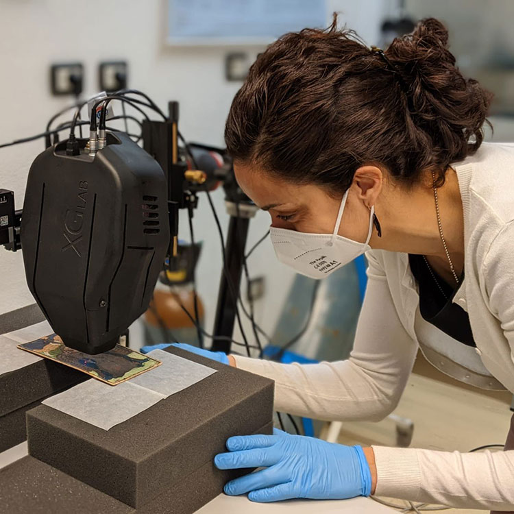 A person in a white FFP2 mask and blue gloves leans over a small, colorful object, examining it under a black scientific device labeled 'XGLAB'. The object rests on white paper within black foam supports.