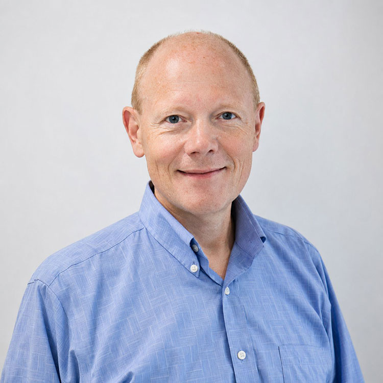 A man with a bald head and blue eyes smiles, looking forward. He wears a light blue button-down shirt.