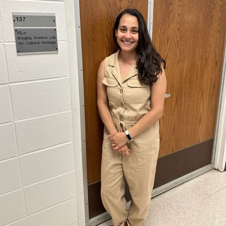 A woman with long dark hair, wearing a tan sleeveless jumpsuit, stands smiling next to a wooden door. A sign on the white brick wall reads "137 ISLα Imaging Science Lab for Cultural Heritage."