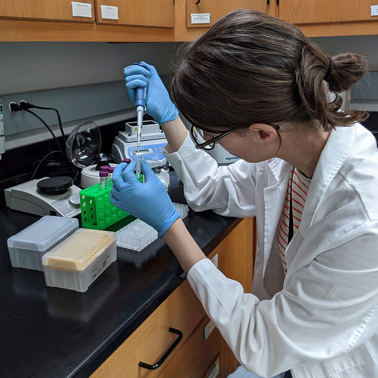 A person in a white lab coat and blue gloves uses a micropipette to transfer liquid between small vials on a dark laboratory bench. Shelves and other lab equipment are visible in the background.