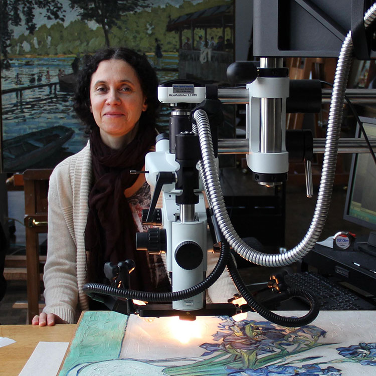 A woman with dark, curly hair smiles at the viewer, seated beside a large scientific microscope. The microscope is positioned over a painting depicting blue and purple flowers, revealing thick brushstrokes. A landscape painting hangs in the background.