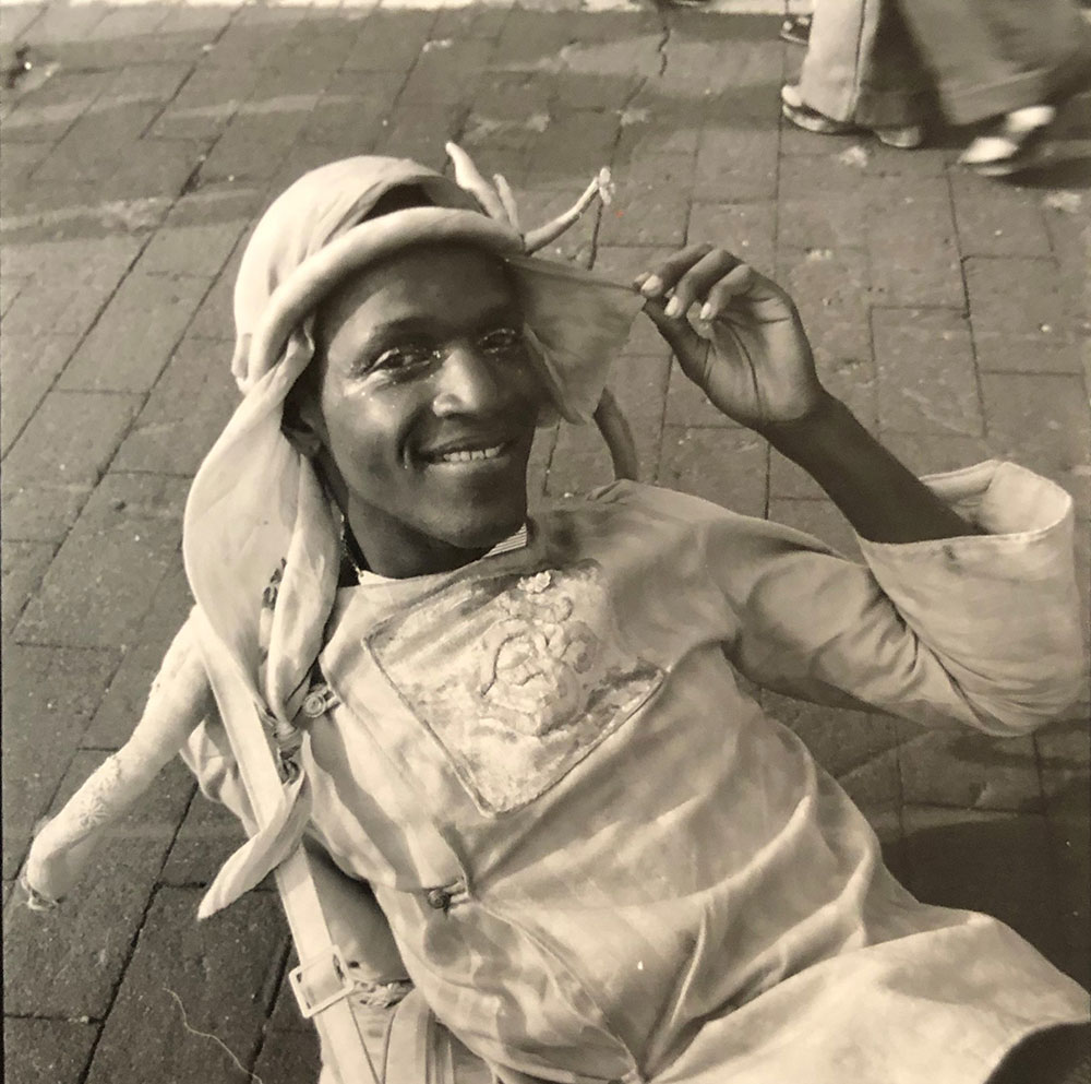 A silver gelatin print photograph with sepia tones of Marsha P. Johnson laying down on a stone path, visible from the waist up. She is wearing a white blouse with three quarter length sleeves and a white keffiyeh on her head. She has one arm up holding a piece of the hat’s fabric aside from her face.