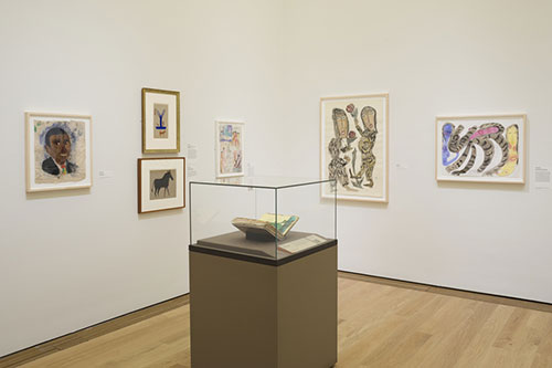 Interior of gallery with white walls, wood floor, a pedastal in the center with sketchbook in disoplay case and five drawings hanging on the wall.