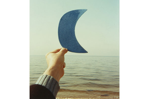 Color photograph of a hand holding up a crescent moon shaped object that is blue with the sea and sky in the background.