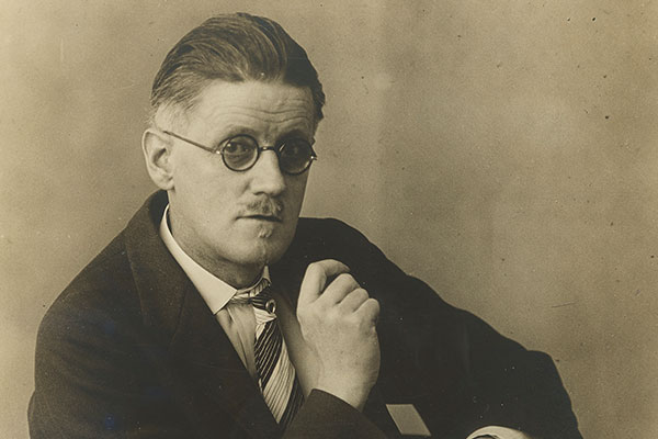 Sepia toned photograph of James Joyce seated looking at camera wearing glasses.
