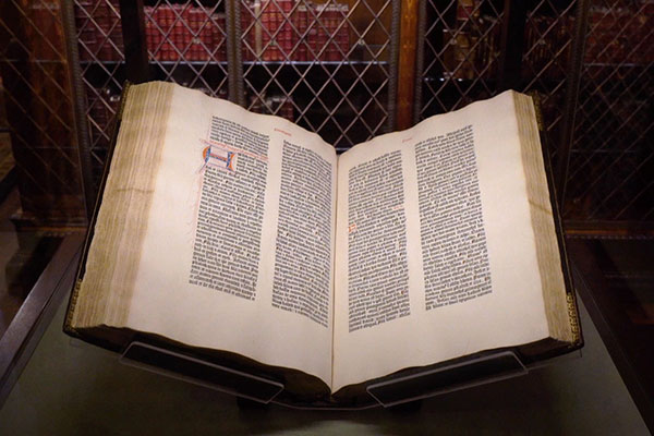 Open Gutenberg Bible displayed in case with bookshelves in the background.