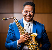 Steven Banks holding a saxophone wearing a blue suit, tie, and glasses