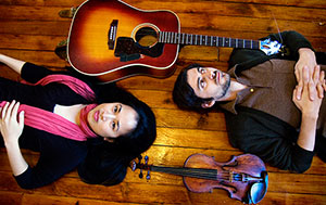 Male and female figures laying on wooden floor looking up at camera from above with guitar laying on one side and violin on the other.