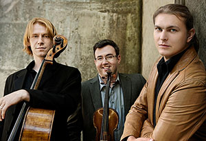 Three men looking forward, the one of the left holds a cello.