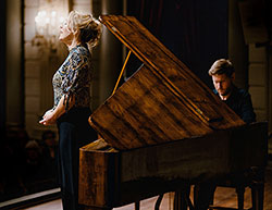 Woman facing left singing while man is playing piano at right.