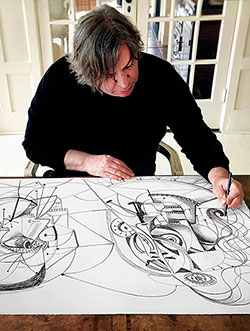 Overhead view of George Condo sitting at table wearing a black top drawing in black pen opn a large white sheet.