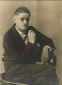 Sepia toned photograph of James Joyce seated with legs crossed and wearing glasses.