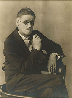 Sepia toned photograph of James Joyce seated looking at camera wearing glasses.