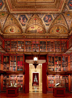 Interior photograph of J. Pierpont Morgan's Library showing ornate gold ceiling with paintings, three tier bookshelves and doorway to Rotunda.