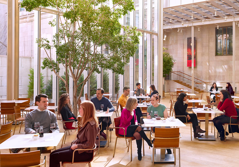 Sunlit view of cafe with tabl;es and chairs and visitors sitting enclosed in a glass atrium.