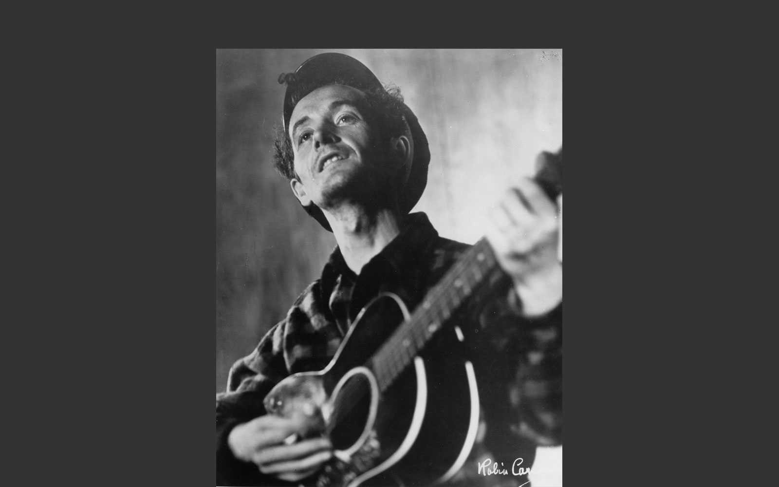Black and white photo of Woody Guthrie playing guitar wearing cap and checked shirt.