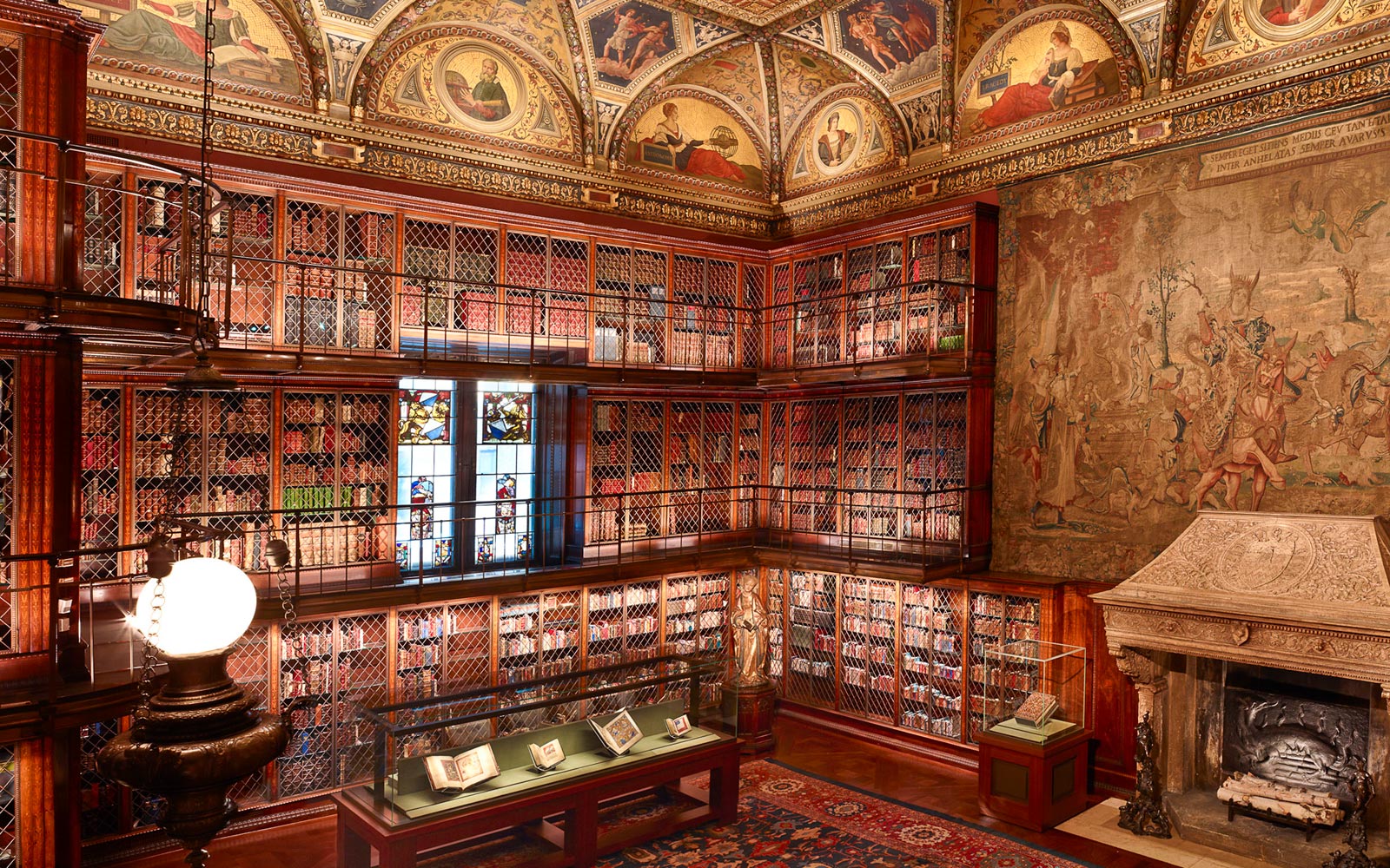 Interior space of J. Pierpont Morgan’s Library with ornate muraled gold ceiling, bookshelves, a tapestry hanging on the wall over a fireplace, and display cases on the floor containing books.