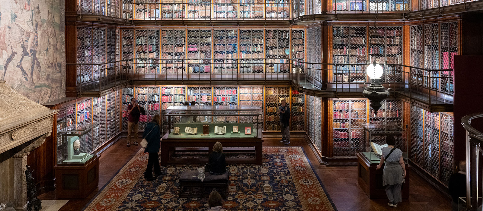 Interior space of J. Pierpont Morgan’s Library with ornate muraled gold ceiling, bookshelves, a tapestry hanging on the wall over a fireplace, and display cases on the floor containing books.