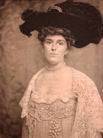 A sepia tinted portrait of a woman looking directly at the camera, visible from the waist up in front of a plain background. She wears a white lace loose bodice dress with a square neck, embroidered detail and draped lace sleeves, a white pearl necklace that sits tightly around her neck, a large dark hat with an equally large plume of the same color, and her hair in an updo.