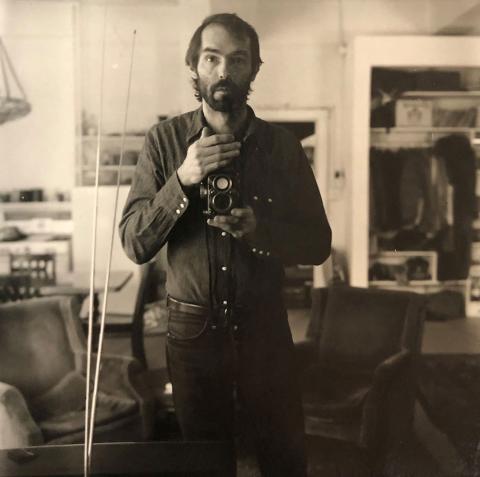 A silver gelatin print photograph with sepia tones of Peter Hujar standing in his apartment, visible from the knees up. He is holding a rolleiflex camera in one hand at the center of his chest with his other hand covering the pop up viewfinder. He is looking up from the camera and forward in the mirror. He wears a dark long sleeve pearl snap shirt tucked into black jeans with a belt. The background apartment is out of focus, but two velvet seats, some shelves, and a closet are visible.