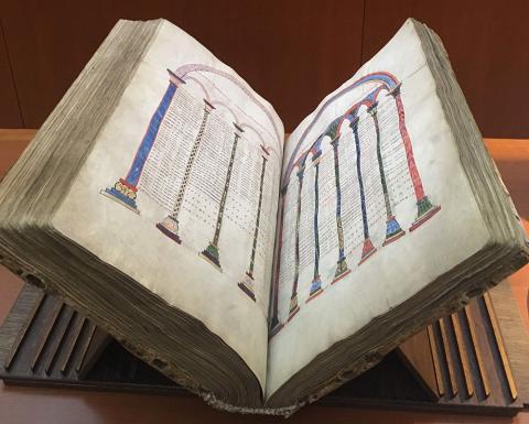 A large bound manuscript sitting open in a brown wooden book stand on a brown wooden table. The opening shows illustrations consisting of different colored pillars connected by arches at the top, with writing between the pillars.