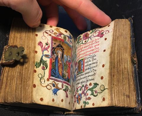 A small and thick bound Book of Hours sitting on a blue velvet pillow held open by a human hand, with a brown leather and gold metal clasp hanging over the pages of the manuscript on the left. The left side opening shows a colored miniature with a floral border. The right side opening has handwritten text in black and red and a decorated O initial, all surrounded by the same floral border.