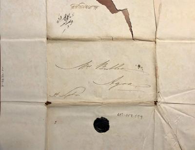 A yellowing piece of unfolded paper with creased lines that indicate it was once folded, with an address and name handwritten in a faded black ink in the center above a black wax seal. Above the address, at the top in the is a tear in the paper, where a brown wooden table is visible underneath.