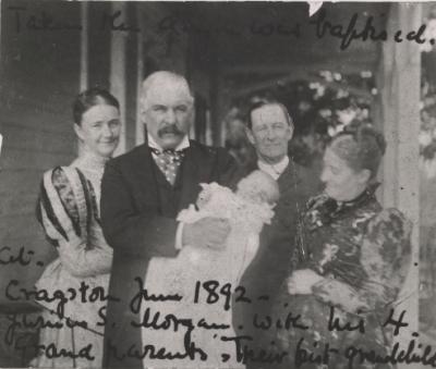 A black and white photograph featuring J. Pierpont Morgan holding a baby with two women and a man surrounding him. The baby is dressed in a white gown while Morgan and the other man wear black suits and the women wear dresses, both with long puffed sleeves and high necks. There are some handwritten notes in black scribbled on the photograph, the most legible reads 1892.