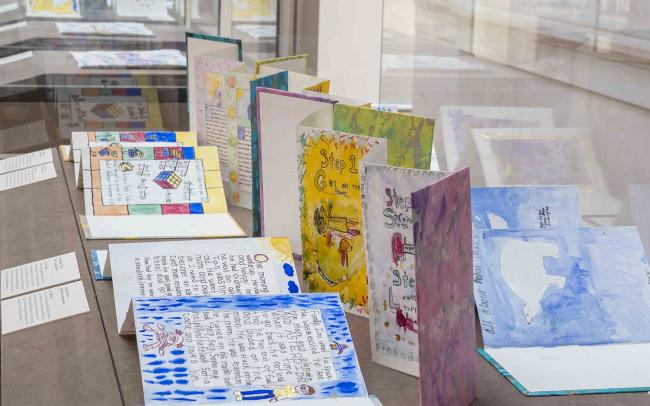 Photograph of exhibition case containing books created by school students with blue and yellow cover with designs and text painted on them.