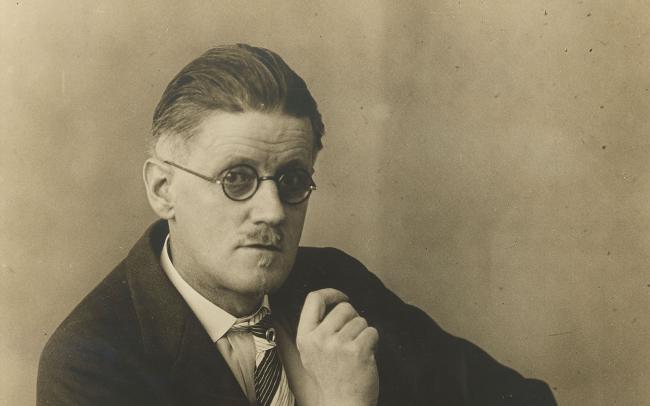 Sepia toned photograph of James Joyce seated looking at camera wearing glasses.