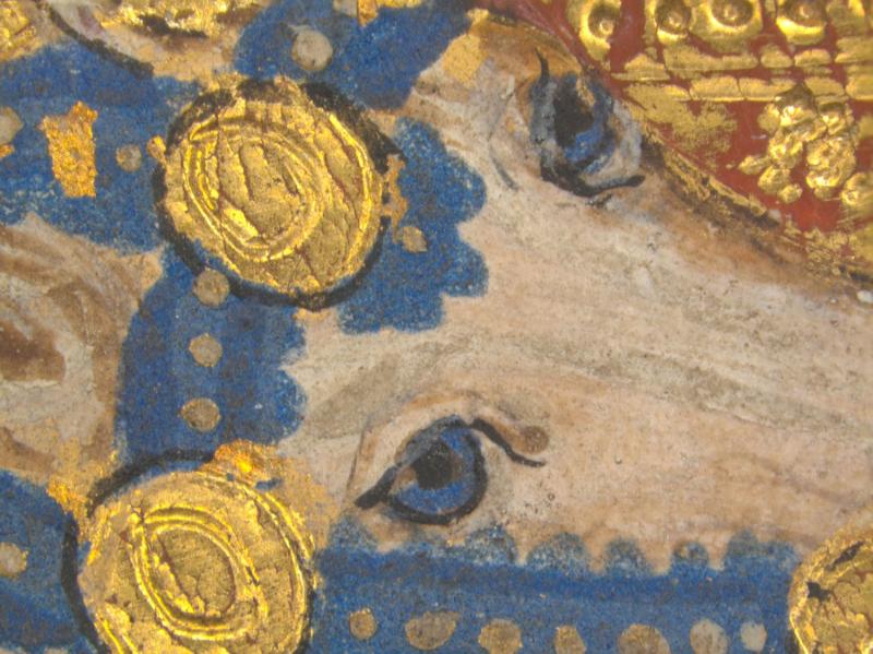 A close-up of a painted face, possibly an animal, with a blue eye and black pupil. The pale face is surrounded by textured blue pigment and raised gold leaf, some in circular patterns, with red pigment visible in the upper right.