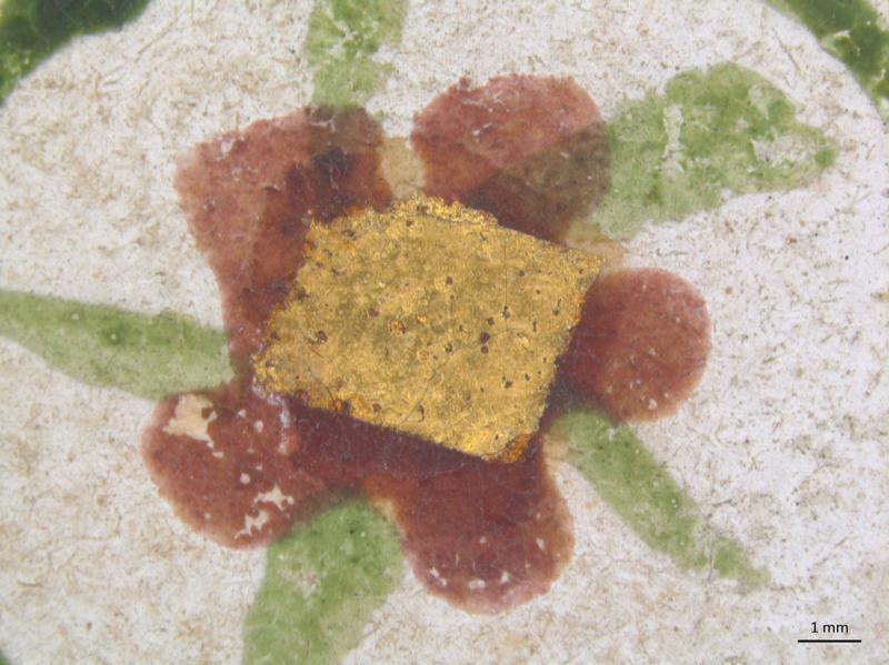 A close-up shows a rectangular piece of gold leaf centered on a textured, light-colored surface. Surrounding the gold leaf are painted reddish-brown and green organic shapes. A 1 mm scale bar is visible in the bottom right.