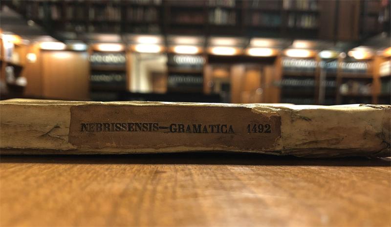 The spine of a book that reads Nebrissensis-Gramatica 1492 in black type on a brown piece of parchment pasted onto a light beige parchment that makes up the binding. The shot is focused on the spine of the book that lays flat on a brown wooden table with the background blurred.