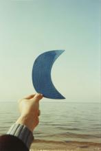 Color photograph of a hand holding up a blue baseball cap bill with the sea and sky in the background.