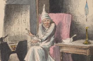 Seaetd figure of Scrooge in white night gown and cap sitting in pink armchair next to a fireplace on the left with a table and lamp on the right.